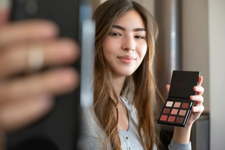 A young woman live streaming a makeup tutorial from her home office, using a smartphone.