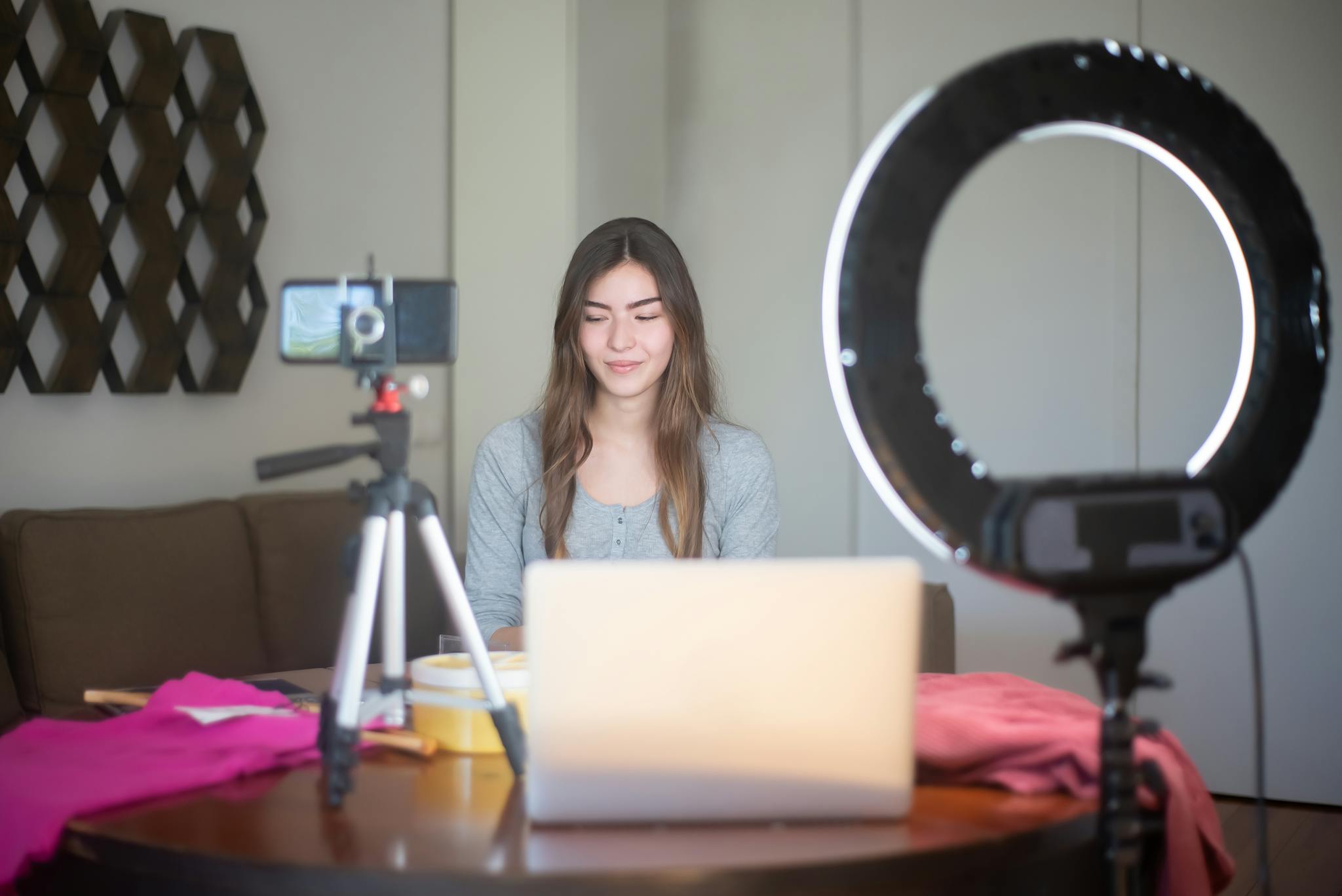 A young woman vlogging indoors using a smartphone, tripod, and ring light setup.